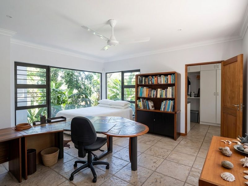 A spacious bedroom featuring large corner windows with garden views, tiled flooring, a ceiling fan, and a built-in wardrobe.