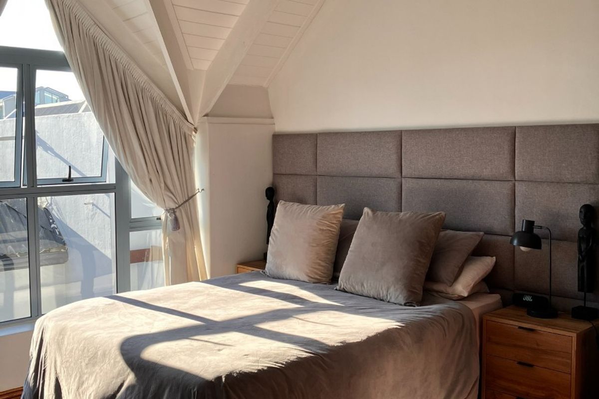 A well-lit bedroom featuring a vaulted ceiling with exposed timber beams, large casement windows, and a modern upholstered headboard.