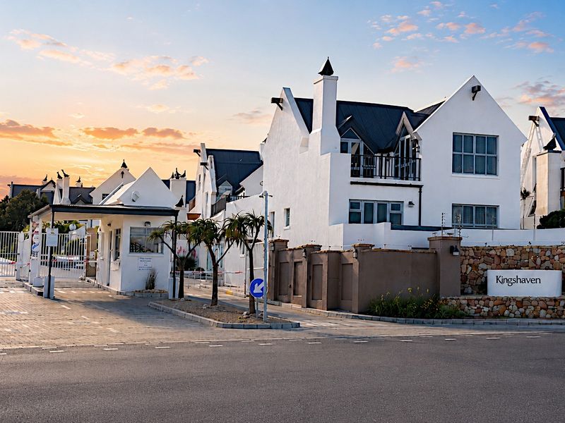 The entrance to Kingshaven townhouse complex featuring a secure gatehouse, paved driveway, and well-maintained white facades with stone accents at sunset.