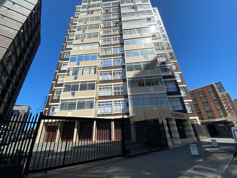 A low-angle exterior view of a multi-story apartment building featuring a secure gated entrance, electric fencing, and multiple balconies with sliding windows.