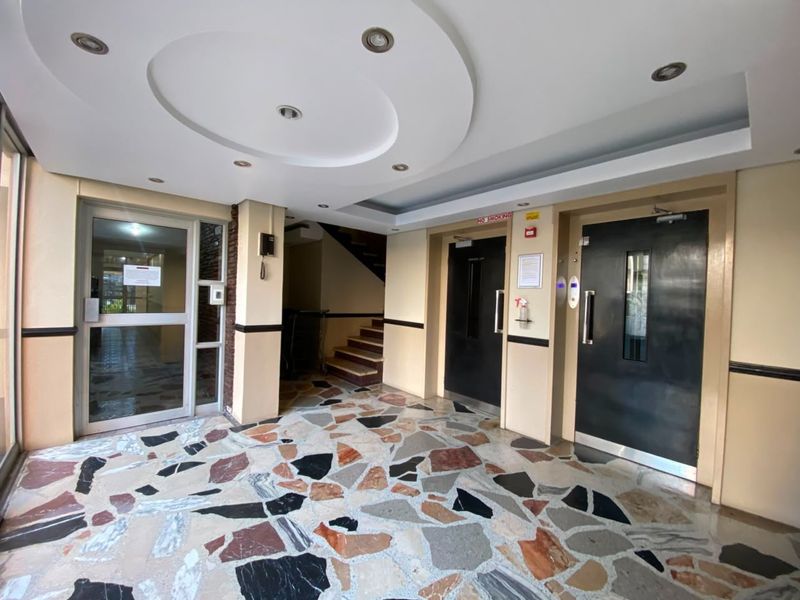 A well-maintained apartment building lobby featuring terrazzo flooring, recessed ceiling lights, elevator access, and a staircase leading to upper levels.