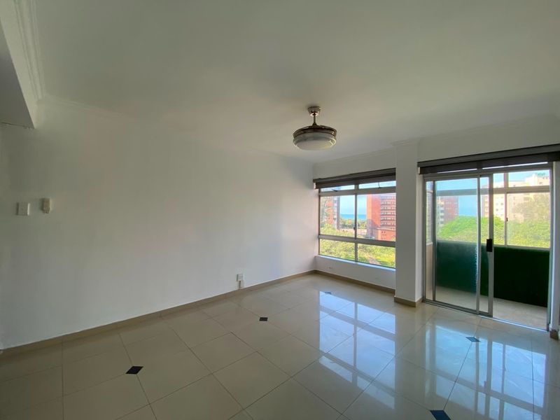 A bright, empty living area featuring tiled floors, a ceiling fan, and large windows with access to an adjoining balcony.