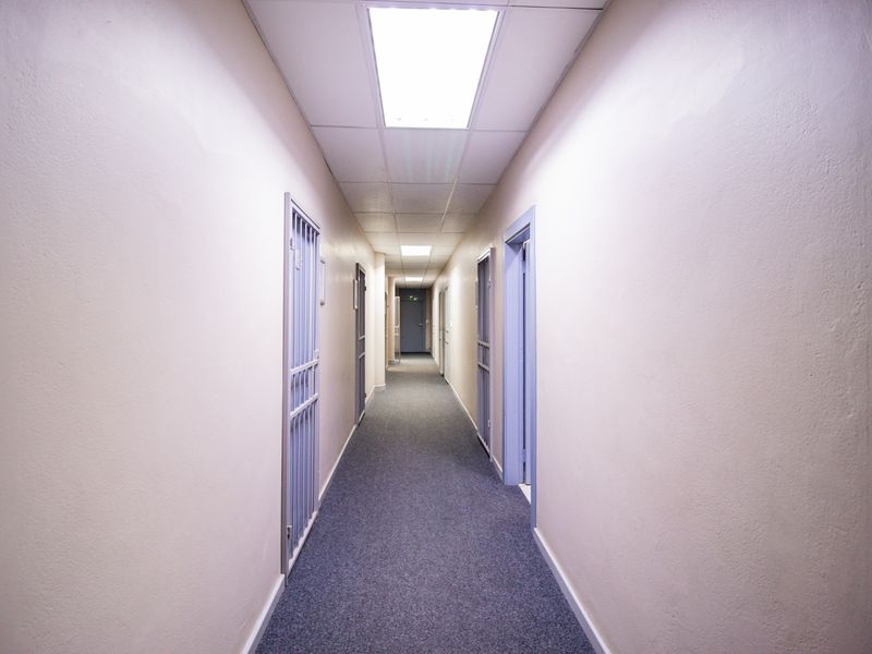 A clean, functional commercial hallway featuring carpeted flooring, suspended ceiling tiles with recessed lighting, and multiple security gates leading to individual office spaces.