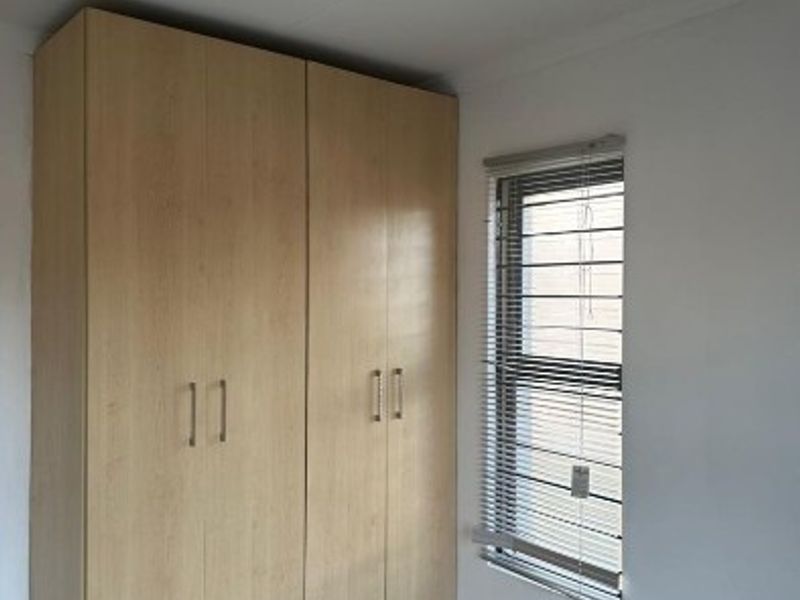 A clean, empty bedroom featuring light wood-finish built-in cupboards, tiled flooring, and a window fitted with security bars and venetian blinds.