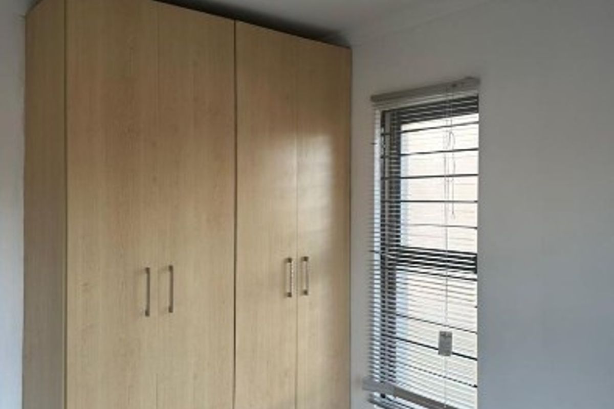 A clean, empty bedroom featuring light wood-finish built-in cupboards, tiled flooring, and a window fitted with security bars and venetian blinds.