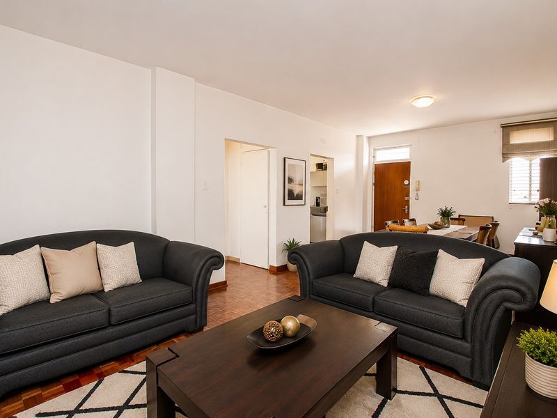 A well-maintained living room featuring classic parquet flooring, neutral walls, and direct access to the entrance and adjacent kitchen area.
