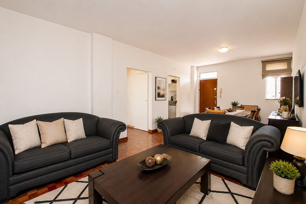 A well-maintained living room featuring classic parquet flooring, neutral walls, and direct access to the entrance and adjacent kitchen area.