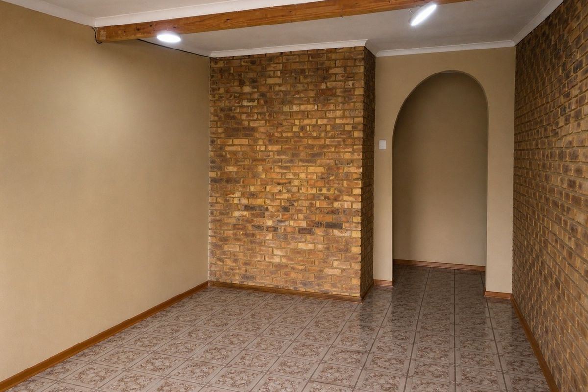 An empty living area featuring an exposed brick accent wall, arched entryway, ceramic tiled flooring, and a decorative timber ceiling beam.