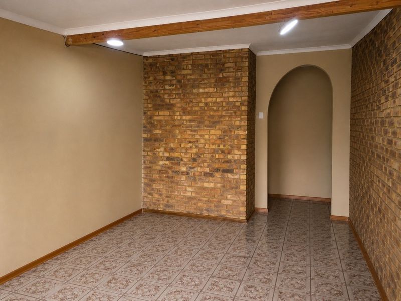 An empty living area featuring an exposed brick accent wall, arched entryway, ceramic tiled flooring, and a decorative timber ceiling beam.