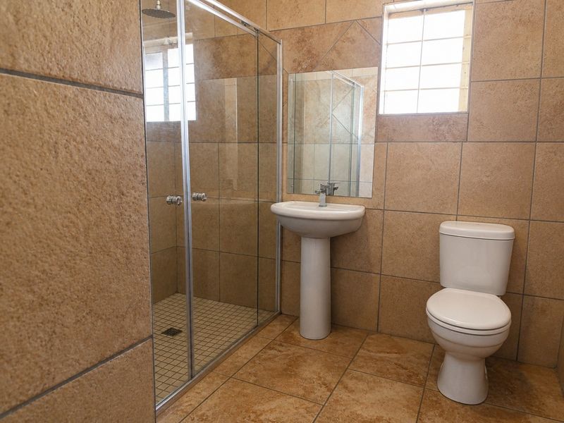 A functional bathroom featuring a glass-enclosed shower, pedestal basin, and toilet, finished with neutral-toned floor and wall tiles.