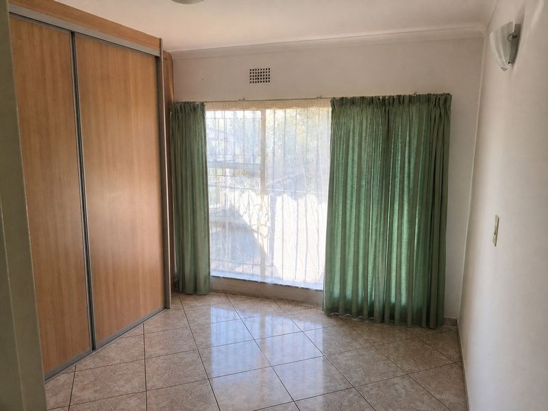 A bright, empty bedroom featuring tiled flooring, a large built-in sliding door wardrobe, and a window fitted with security bars.