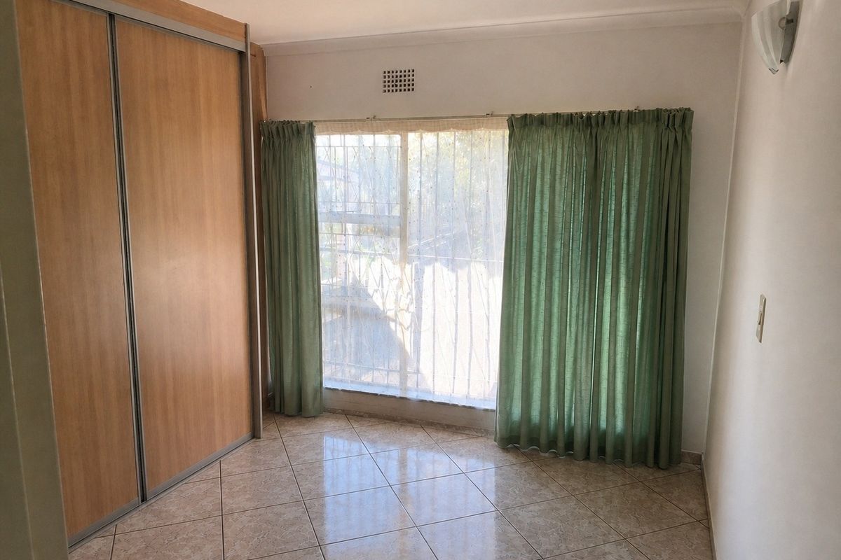 A bright, empty bedroom featuring tiled flooring, a large built-in sliding door wardrobe, and a window fitted with security bars.