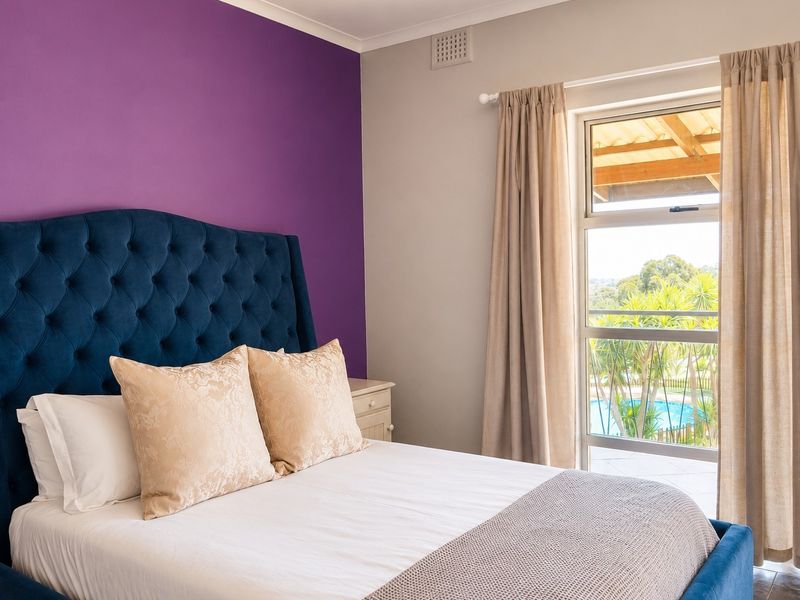 A well-lit bedroom featuring a large window with views of the pool area and garden, finished with neutral and bold accent wall paint.
