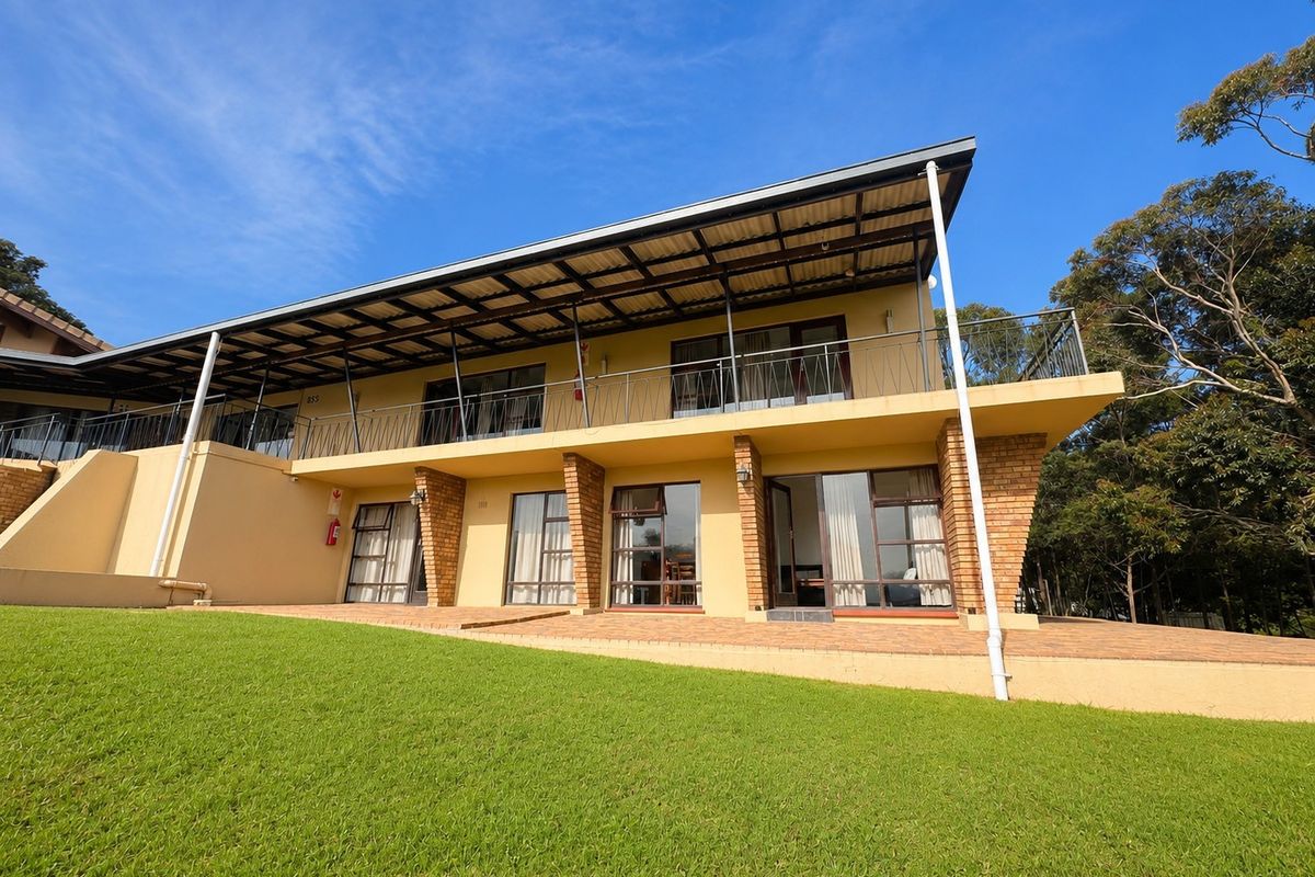 Rear view of a two-story residential building featuring a covered balcony, paved patio area, and brick structural pillars set against a landscaped lawn.