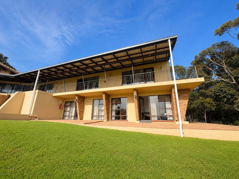 Rear view of a two-story residential building featuring a covered balcony, paved patio area, and brick structural pillars set against a landscaped lawn.