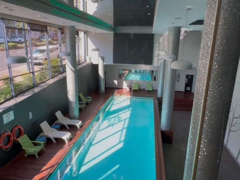 A modern indoor swimming pool area featuring mosaic-tiled columns, timber decking, and expansive floor-to-ceiling windows providing natural light.