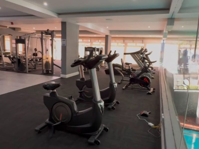 A well-lit, modern apartment gym facility featuring rubber flooring, recessed lighting, and a glass balustrade overlooking the pool area.