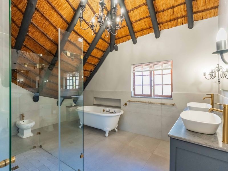 A luxurious bathroom featuring a freestanding bathtub, double vanity with brass fixtures, glass shower enclosure, and a unique exposed thatch roof with timber beams.