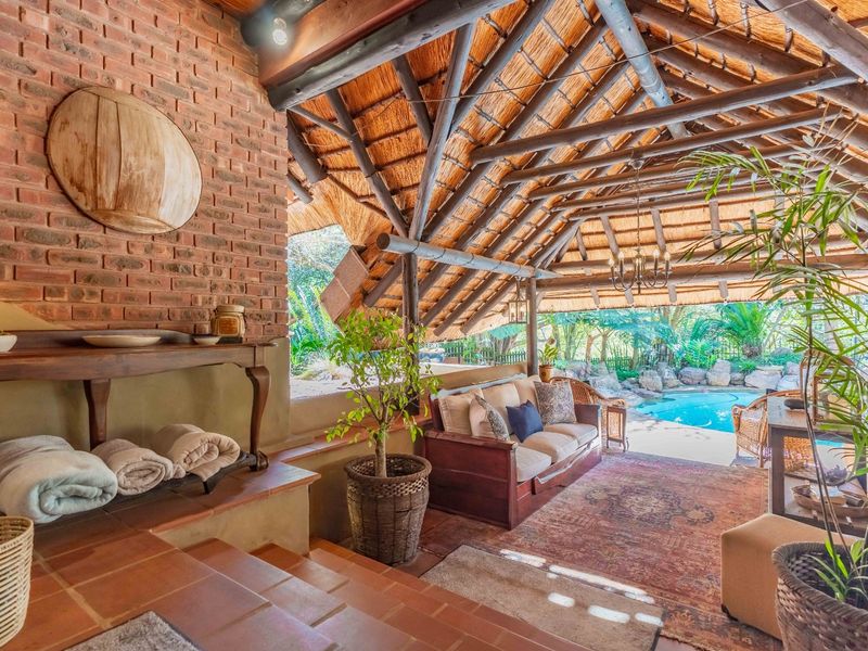 A rustic covered patio featuring a thatched roof with exposed timber beams, exposed brickwork, and terracotta flooring, overlooking a private swimming pool.