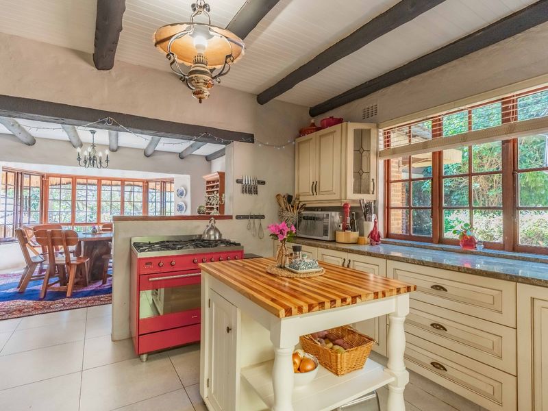 Open plan kitchen featuring exposed timber beams, a central timber island, stone countertops, and a vibrant red gas stove, flowing into the dining area.