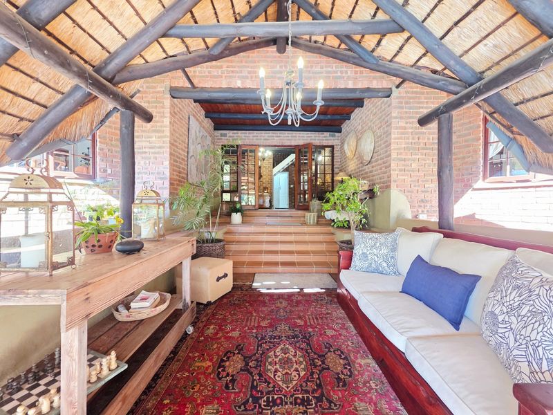 A spacious covered patio featuring a traditional thatched roof, exposed timber beams, and rustic brick walls, leading into the main entrance via tiled steps.