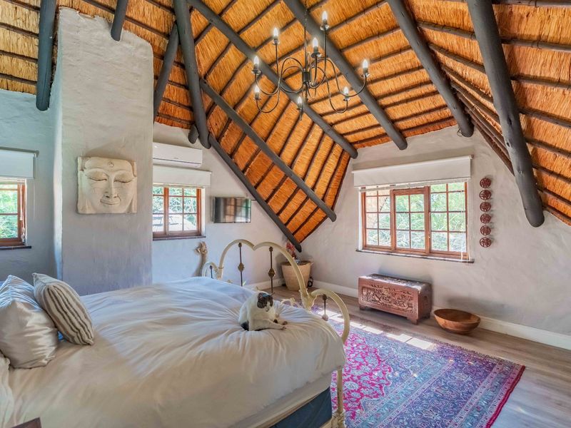 Spacious bedroom featuring a high vaulted ceiling with exposed timber rafters and traditional thatch roofing, complemented by plastered walls and timber-framed windows.