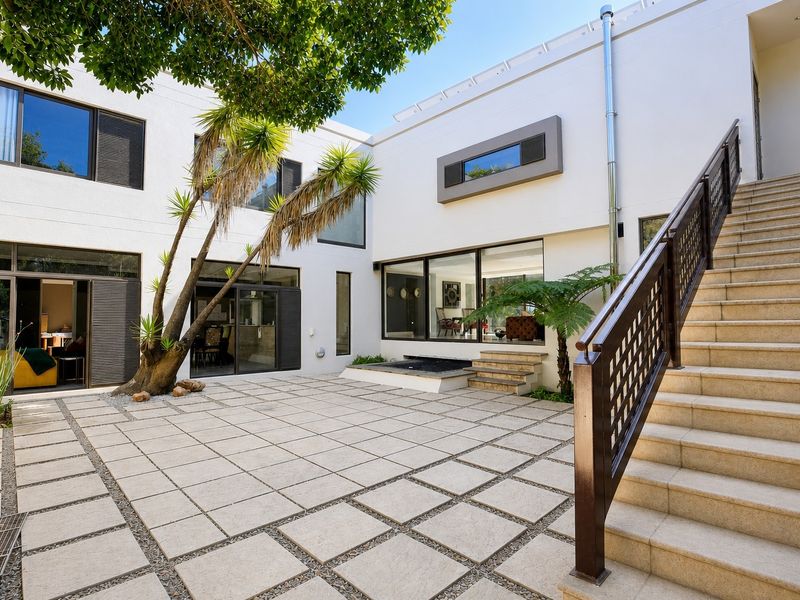 A modern, paved rear courtyard featuring an external staircase with metal balustrades, large sliding glass doors, and a built-in water feature.