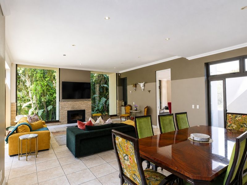 Spacious open-plan living and dining area featuring tiled floors, a stone-clad fireplace, and large windows overlooking lush greenery.