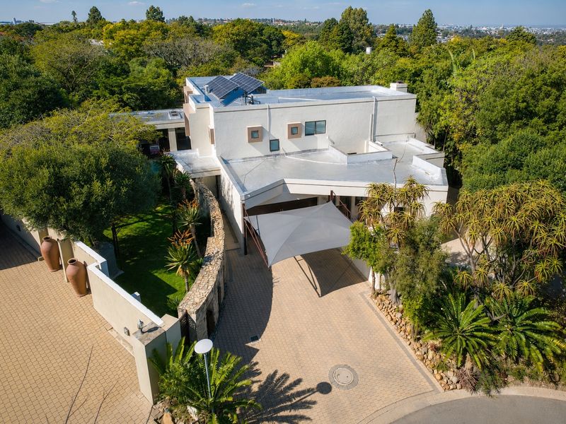 Aerial view of a modern residential property featuring a paved driveway, solar panels, flat roof architecture, and established garden surroundings.