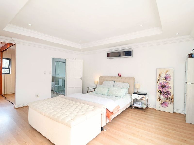 Spacious bedroom featuring a tray ceiling with recessed lighting, light wood-look flooring, a wall-mounted air conditioner, and an adjoining en-suite bathroom.