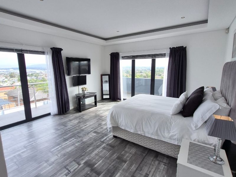 Spacious bedroom featuring modern wood-look tile flooring, recessed tray ceiling lighting, and dual sliding glass doors opening onto a private balcony with scenic views.