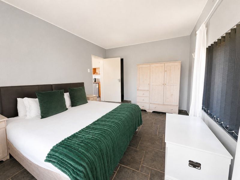 A bedroom featuring tiled flooring and vertical blinds, with an adjacent doorway leading to a kitchen area.
