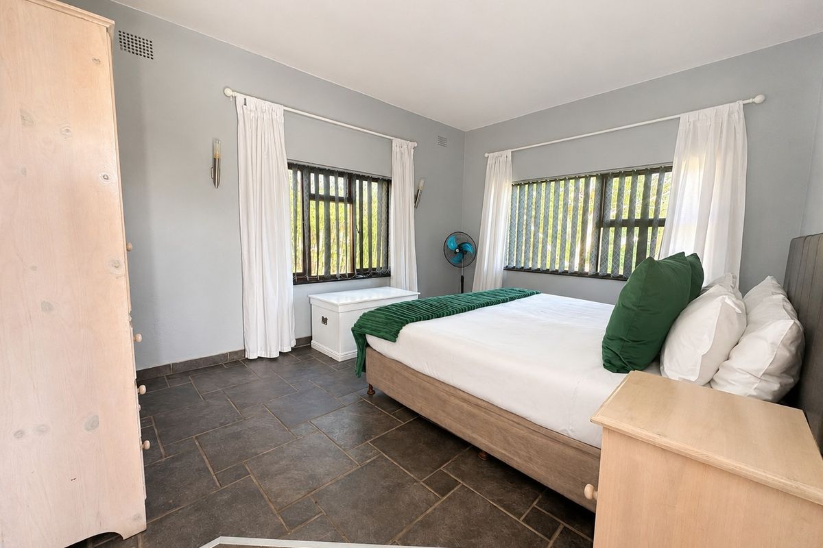 A spacious bedroom featuring dark tiled flooring, neutral wall tones, and dual windows with fitted blinds and curtain rods.
