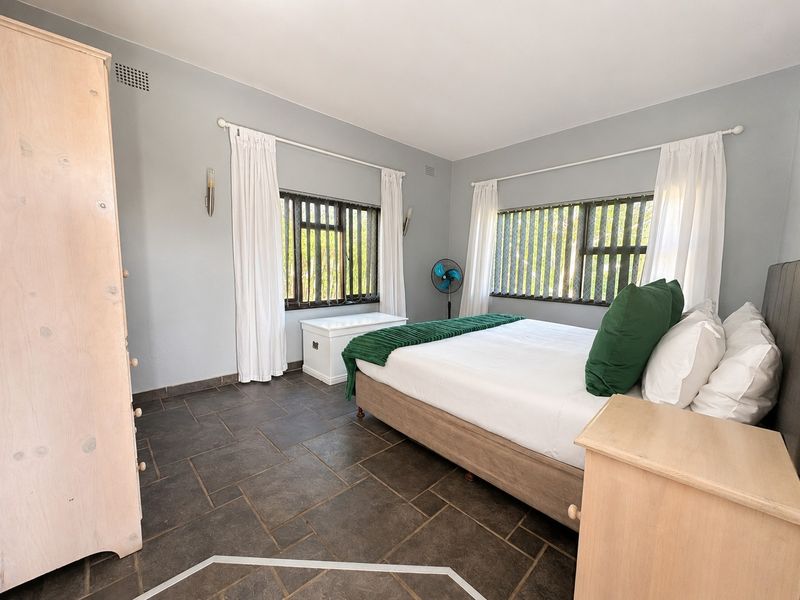 A spacious bedroom featuring dark tiled flooring, neutral wall tones, and dual windows with fitted blinds and curtain rods.