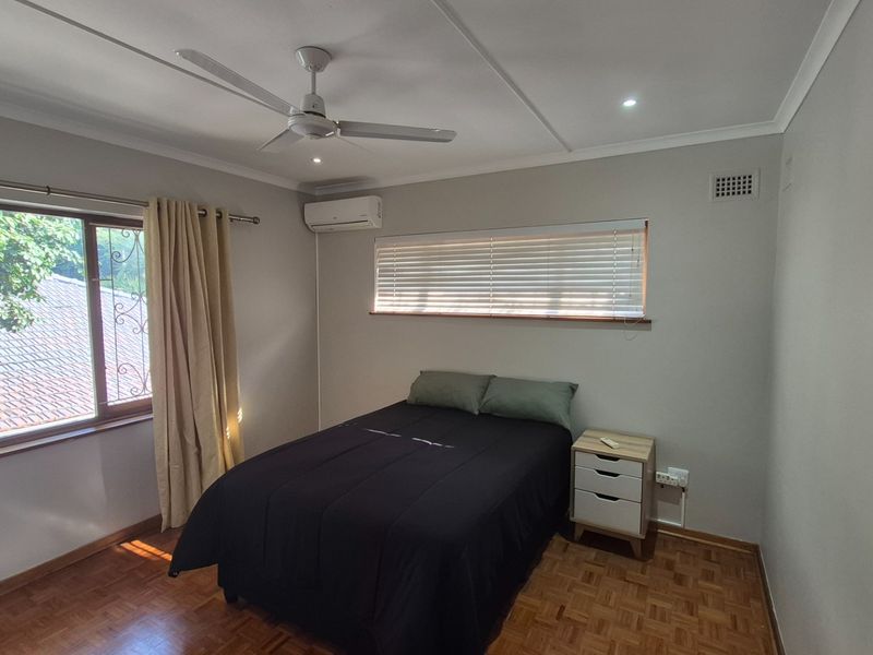 A well-maintained bedroom featuring parquet flooring, a ceiling fan, split-system air conditioning, and large windows with security bars and venetian blinds.