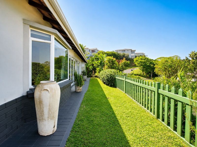 A well-maintained rear exterior view featuring a paved walkway, manicured lawn, and a green picket fence with a clear view of the surrounding landscape.
