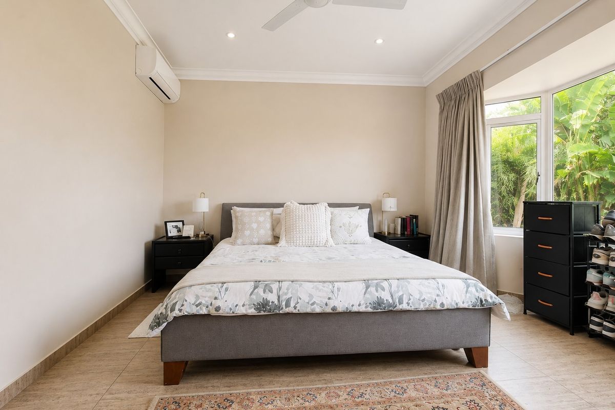 A well-maintained bedroom featuring tiled flooring, a ceiling fan, split-system air conditioning, and a large bay window overlooking the garden.