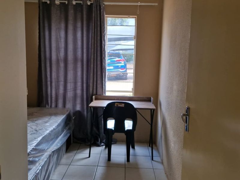 A compact bedroom featuring tiled flooring and a window fitted with security bars, offering a functional space for a single bed and desk area.