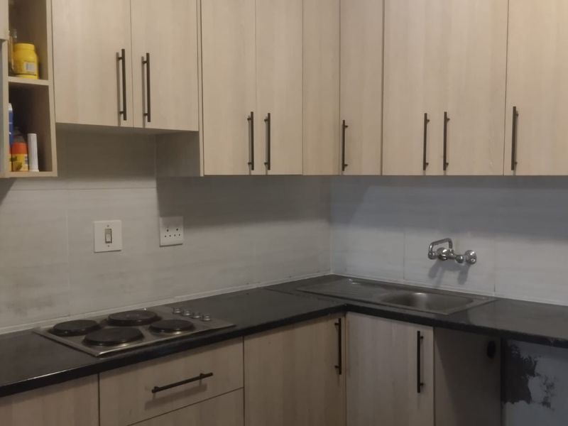 A functional kitchen featuring light wood-finish cabinetry, a dark countertop, an integrated electric hob, and a stainless steel sink with a tiled backsplash.