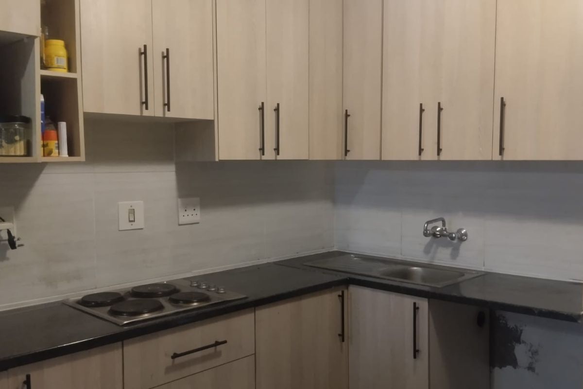 A functional kitchen featuring light wood-finish cabinetry, a dark countertop, an integrated electric hob, and a stainless steel sink with a tiled backsplash.