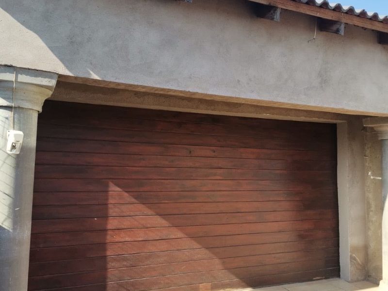 A garage exterior featuring a dark wood sectional door, tiled entrance area, and exposed structural columns under a simple roof overhang.