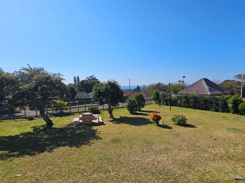 Spacious garden area featuring a well-maintained lawn, perimeter fencing, and a built-in stone braai, offering a pleasant outdoor space with distant sea views.