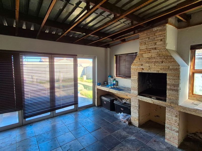 Covered patio area featuring a built-in stone braai, tiled flooring, and exposed timber ceiling, offering direct access to the garden through sliding glass doors.