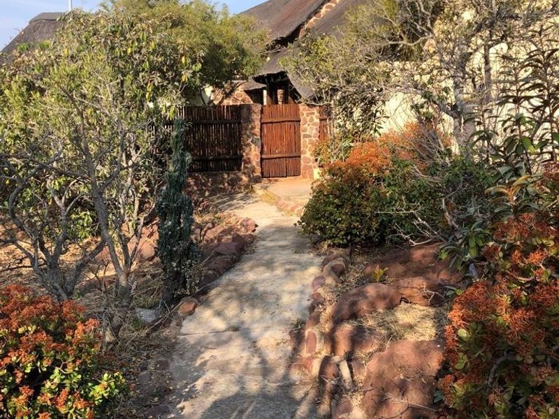 A front entrance path leading to a rustic house featuring traditional thatch roofing, stone masonry walls, and a timber gate surrounded by established garden landscaping.