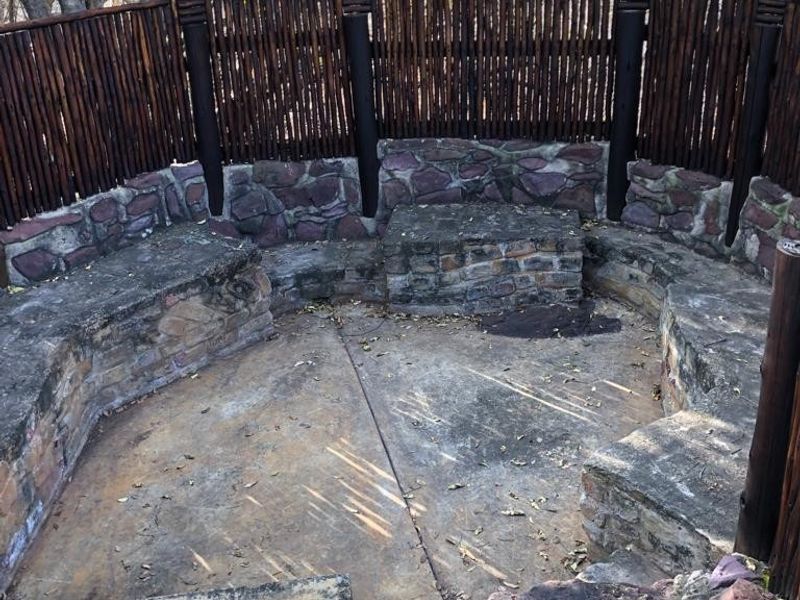 An outdoor boma or patio area featuring built-in stone masonry seating and rustic reed fencing, set in a natural garden environment.