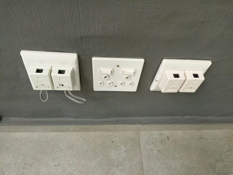 Close-up view of wall-mounted electrical power outlets and data ports installed on a grey textured wall above tiled flooring.