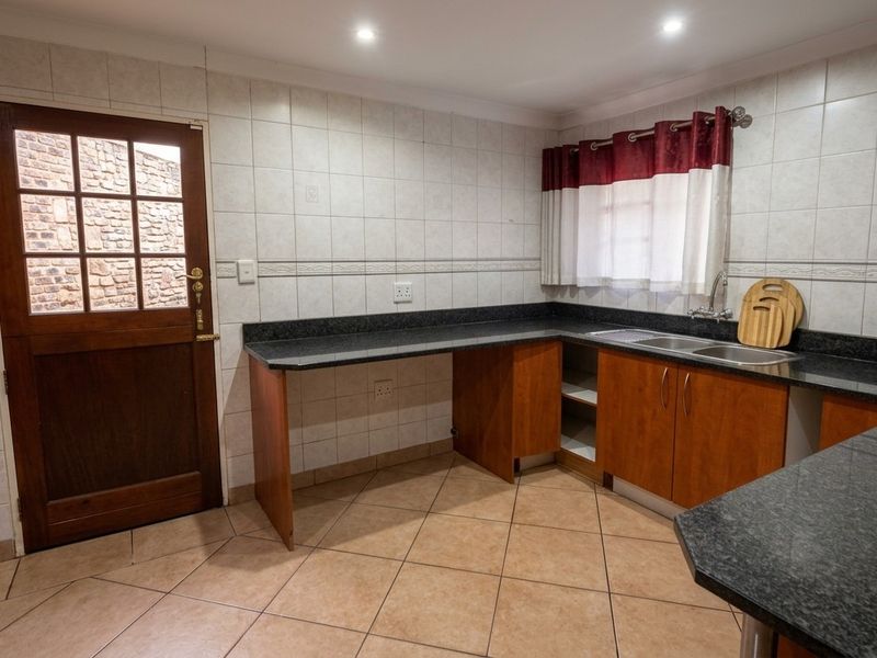 A functional scullery area featuring granite countertops, a double basin sink, tiled finishes, and direct exterior access via a wooden door.