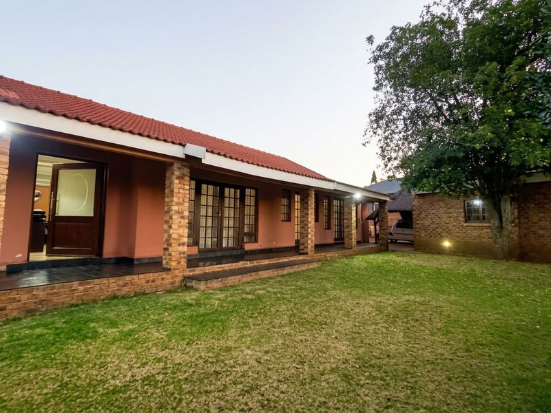 A well-maintained rear exterior view featuring a covered patio with brick pillars, tiled roof, and a spacious lawn area.