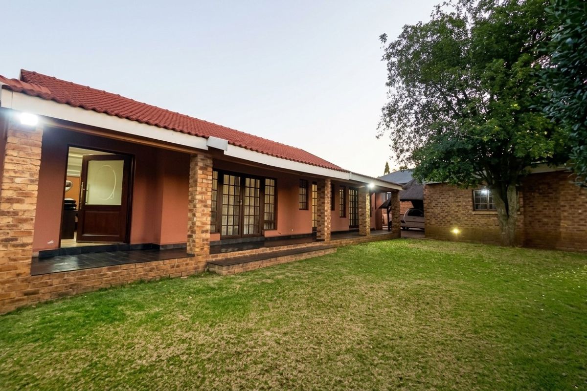 A well-maintained rear exterior view featuring a covered patio with brick pillars, tiled roof, and a spacious lawn area.