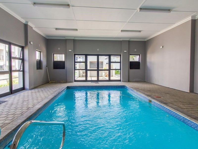 A well-maintained indoor swimming pool area featuring tiled surrounds, large aluminium-framed glass doors, and a clean, neutral interior finish.